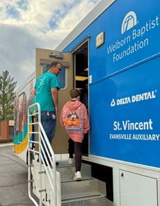 A child with a colorful backpack enters a mobile dental clinic with man opening the door.